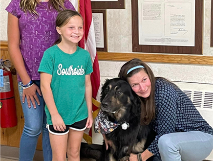 Small girl standing next to a dog while a woman hugs it